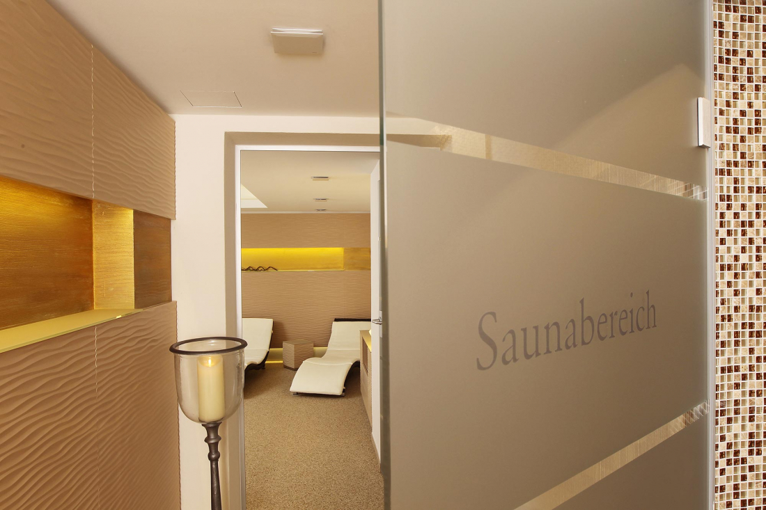 Moderner Wellnessbereich mit beigefarbenen, strukturierten Wänden, warmer Beleuchtung und weißen Liegestühlen. Eine Glastür mit der Aufschrift "Saunabereich" ist teilweise geöffnet und lädt zum Eintritt in den Entspannungsraum ein.
