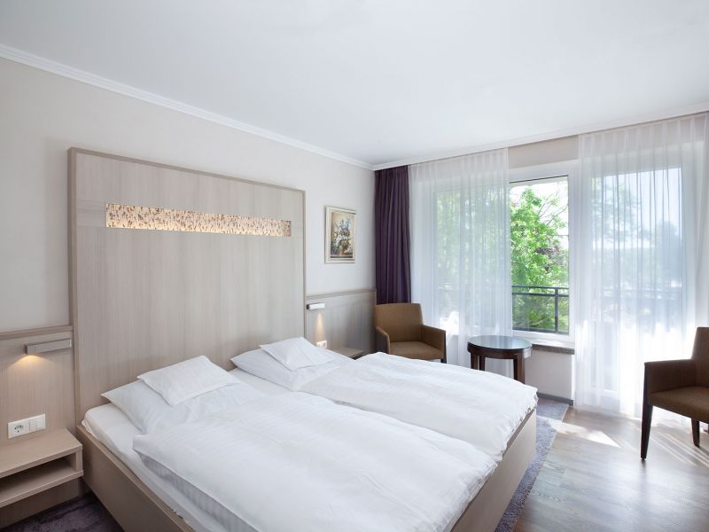 Ein helles, modernes Hotelzimmer mit zwei zusammengeschobenen Einzelbetten, weißem Bettzeug, Holzkopfteil, zwei braunen Stühlen an einem runden Tisch, einem großen Fenster mit durchsichtigen Vorhängen und einem Gemälde an der Wand.