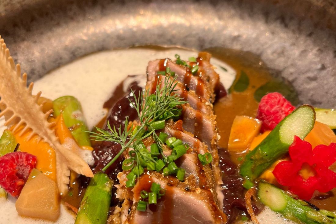 Nahaufnahme eines Gourmetgerichts mit gebratenen Thunfischscheiben und Kräutern, umgeben von Spargel, Karotten, Himbeeren, einer cremigen Sauce und einem dekorativen Knusperstück in einer Steingutschale.