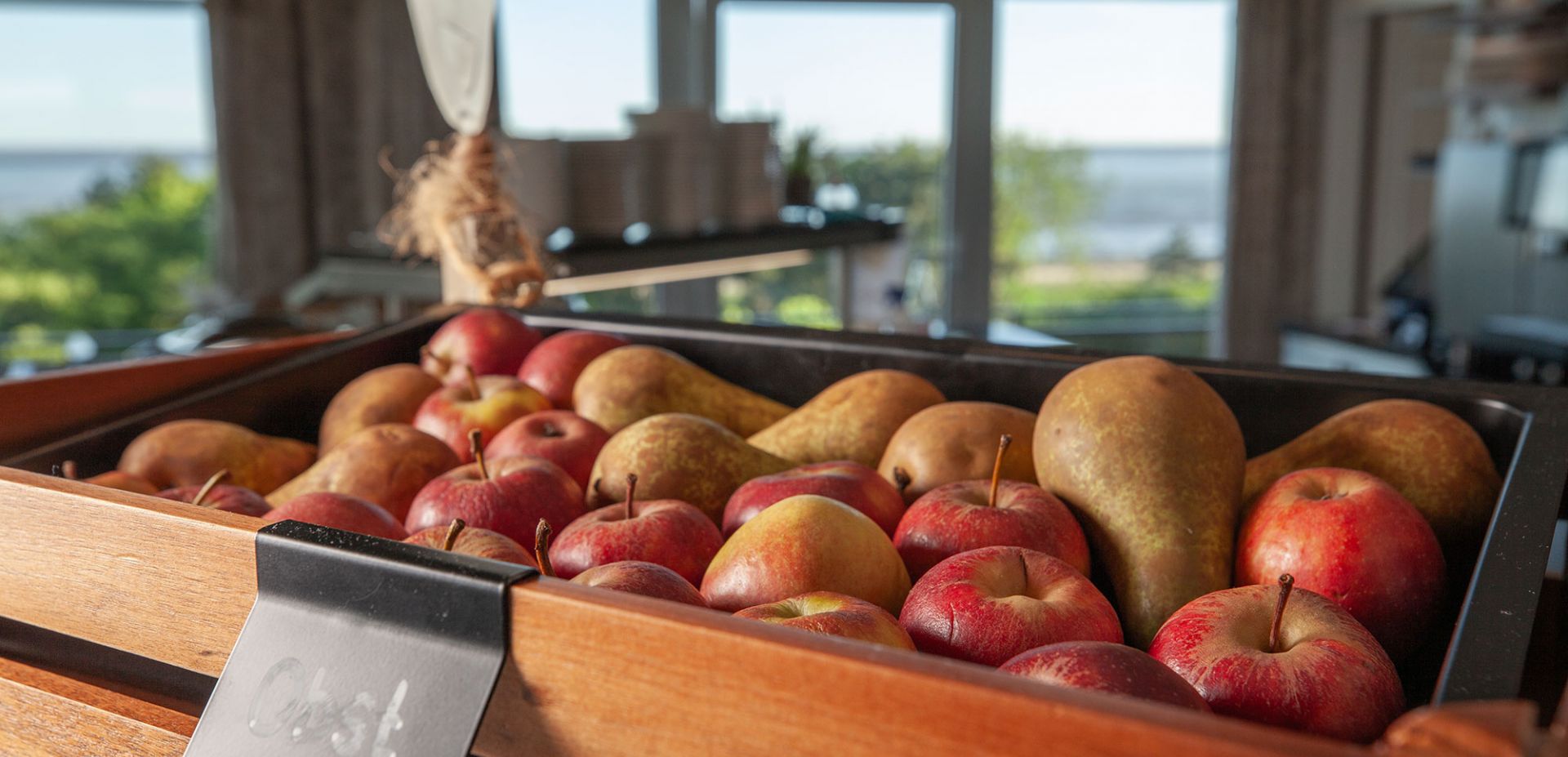 Äfel in einer dunklen Holzkiste mit einem schwarzen Schild auf dem "Obst" steht. Im Hintergund ist eine großzügige Fensterfront mit Blick auf die Nordsee.